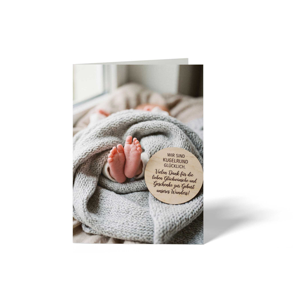 Vorderseite der geschlossenen A6 Dankeskarte zur Geburt von GHG Supplies. Kleine Babyfüße ragen aus einer grauen Strickdecke neben einem runden Holzschild mit dem Text Wir sind kugelrund glücklich. Vielen Dank für die lieben Glückwünsche und Geschenke zur Geburt unseres Wunders!.