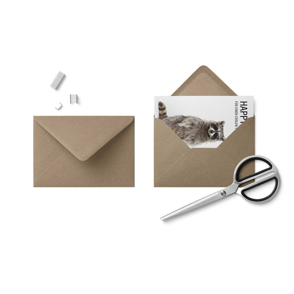 Draufsicht auf das Verpackungsset: Eine geschlossene Geburtstagskarte mit dem Waschbären-Motiv schiebt sich zur Hälfte aus einem braunen, rustikalen Kraftpapier-Briefumschlag mit spitzer Verschlussklappe heraus. Daneben liegen dekorativ platziert eine geschlossene Edelstahlschere und drei kleine Heftklammern auf weißem Hintergrund.