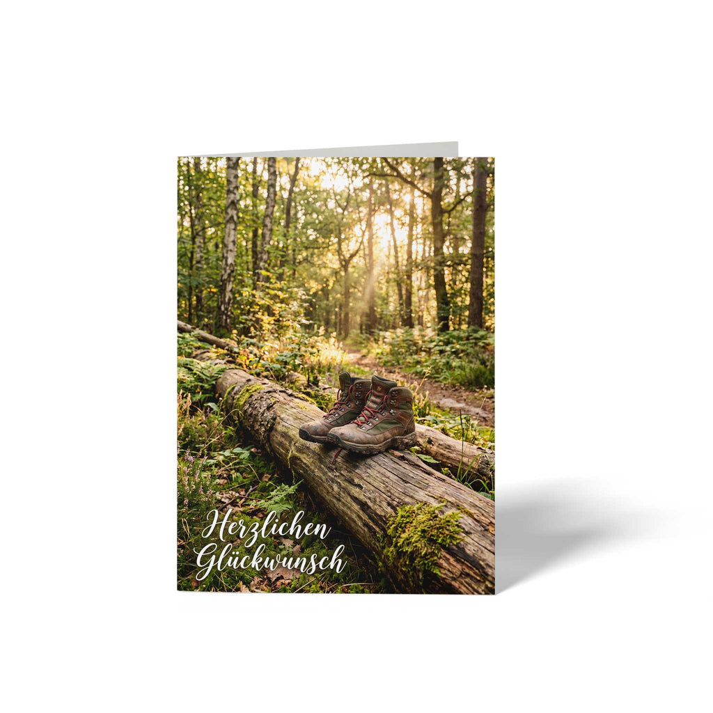 Vorderseite der geschlossenen A6 Glückwunschkarte von GHG Supplies. Ein Paar Wanderschuhe steht auf einem umgestürzten Baumstamm in einem von der Sonne durchfluteten Wald unter dem Text Herzlichen Glückwunsch.