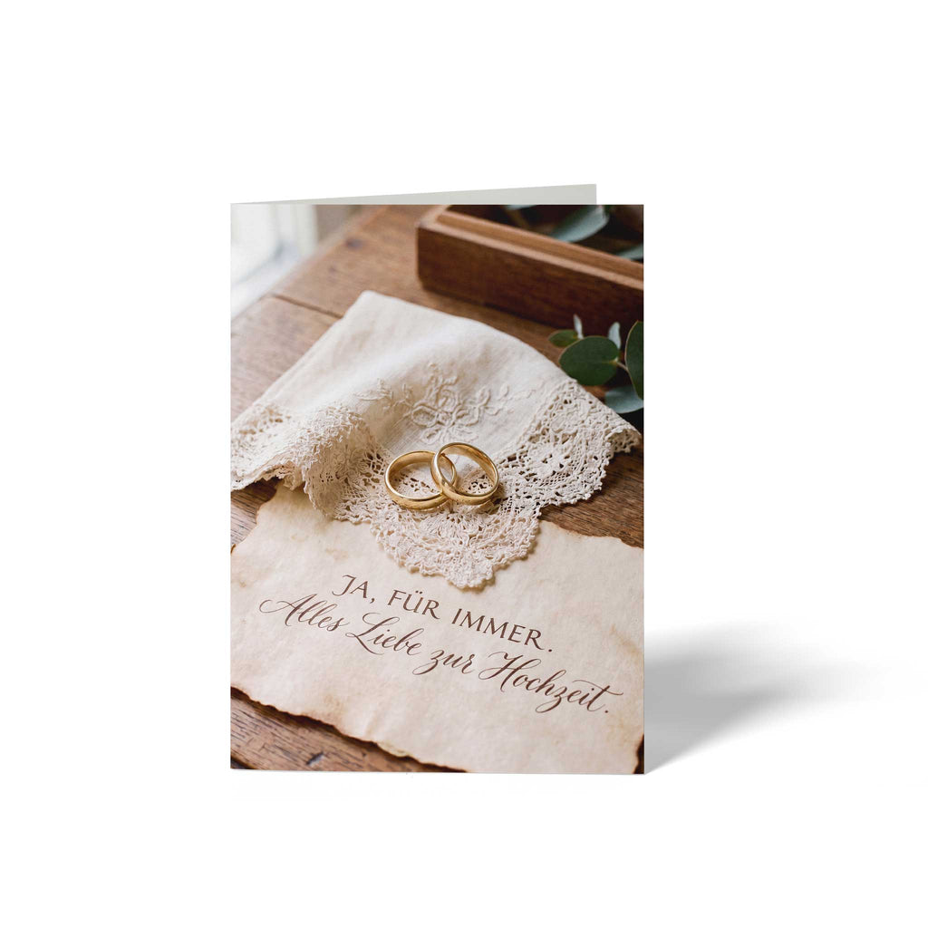 Vorderansicht der geschlossenen A6 Hochzeitskarte von GHG Supplies. Zwei goldene Eheringe liegen auf einem Spitzentaschentuch über Pergamentpapier mit dem eleganten Schriftzug Ja für immer - Alles Liebe zur Hochzeit.