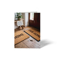 Vorderseite der geschlossenen A6 Hochzeitskarte von GHG Supplies. Zu sehen sind zwei Fußmatten mit den Aufschriften Home und Our Home auf Holzboden sowie ein Zettel mit dem Spruch Ab jetzt wird geteilt. Glückwunsch zur neuen Ära!.