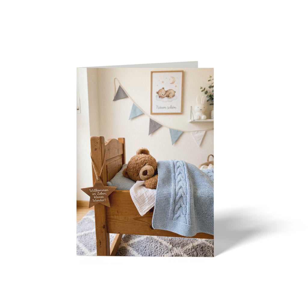 Vorderseite der geschlossenen A6 Glückwunschkarte zur Geburt von GHG Supplies. Schlafender Teddybär im Holzbett unter einer blauen Strickdecke im Kinderzimmer mit Holzstern-Anhänger Willkommen im Leben, kleines Wunder.