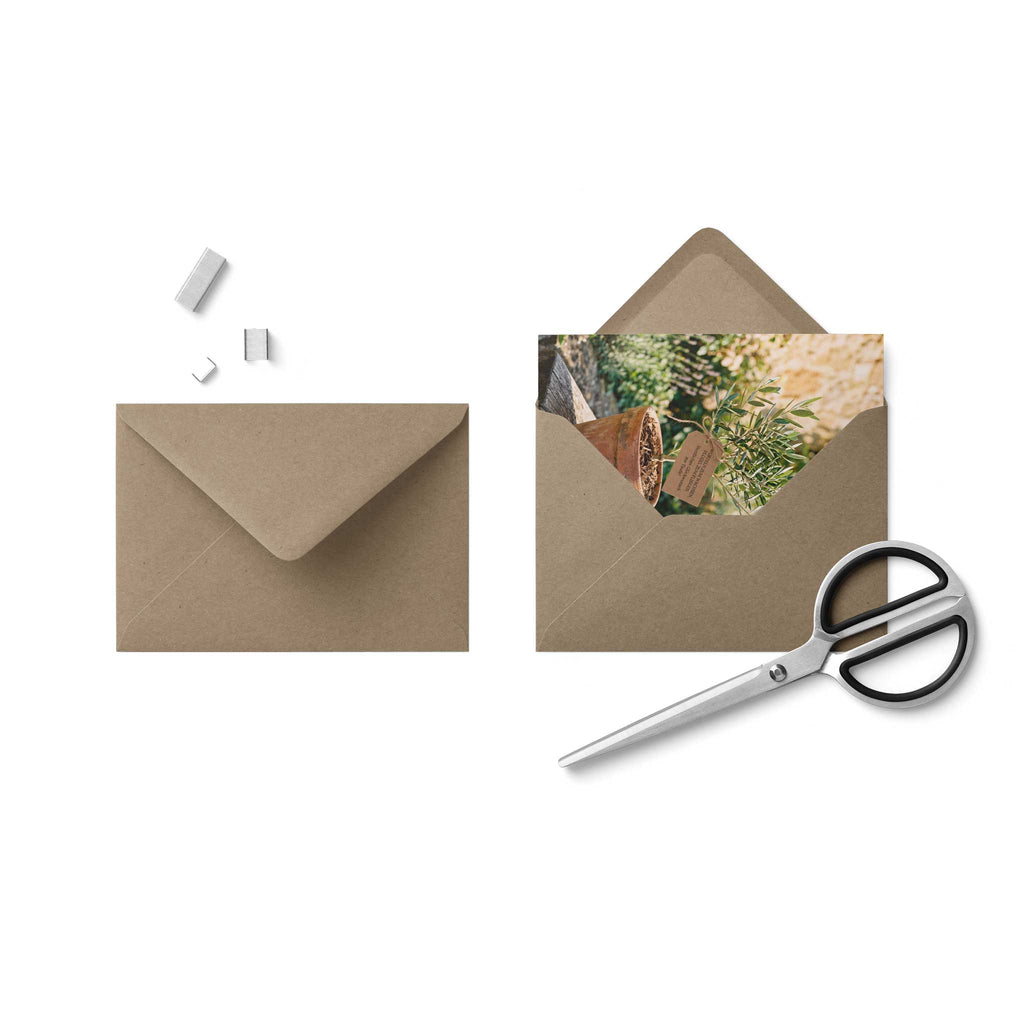 Die hochformatige Faltkarte zur Taufe mit dem Olivenbaum-Motiv liegt leicht versetzt eingelegt in einem passenden, rustikalen braunen Kraftpapier-Umschlag vor weißem Hintergrund neben einer Schere.