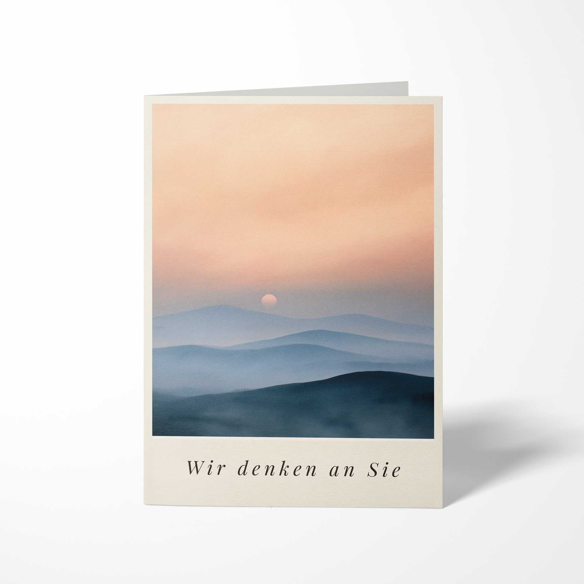 Vorderansicht der hochkant gefalteten B6 Trauerkarte mit einem Aquarell-Motiv blauer Hügelketten im Sonnenuntergang und dem kursiven Schriftzug Wir denken an Sie.
